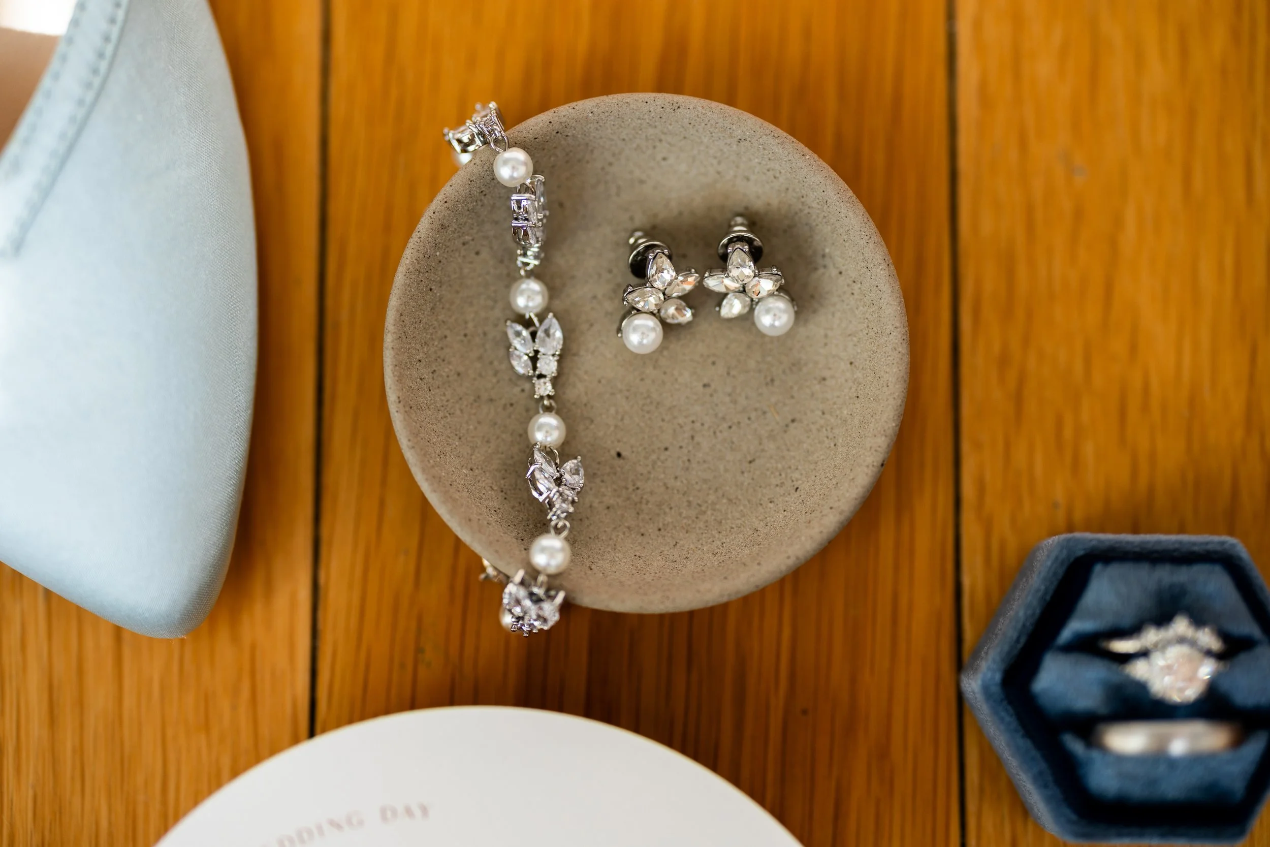 Jewelry set including a pearl and crystal bracelet, matching earrings with pearls and crystals, and a ring with a central gemstone, displayed on a round ceramic dish and in a ring box, on a wooden surface.