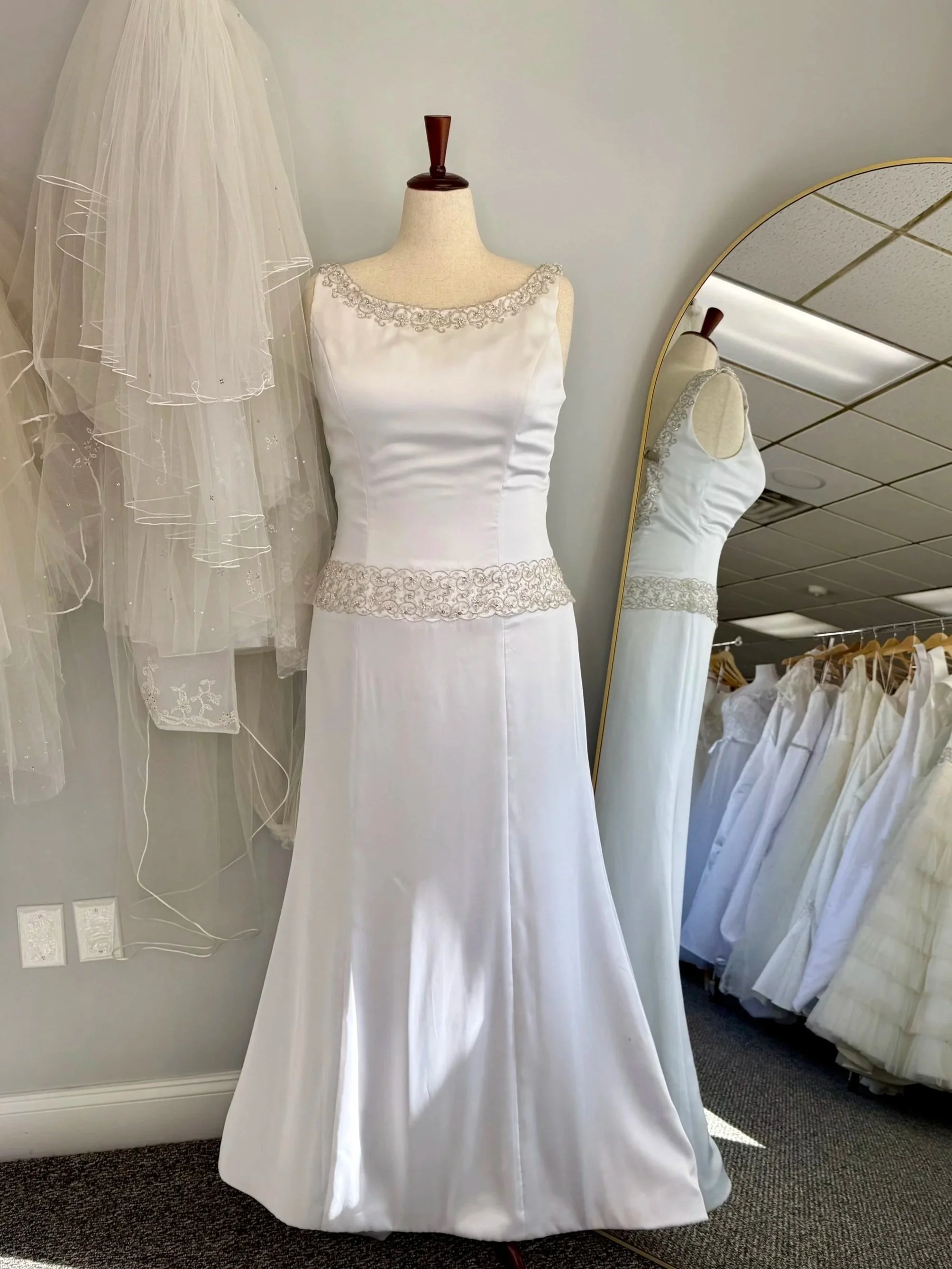 Image of high neck Eden Bridals gown with silver embroidery along neckline and drop waist lineon a mannequin in front of veil wall and store mirror.