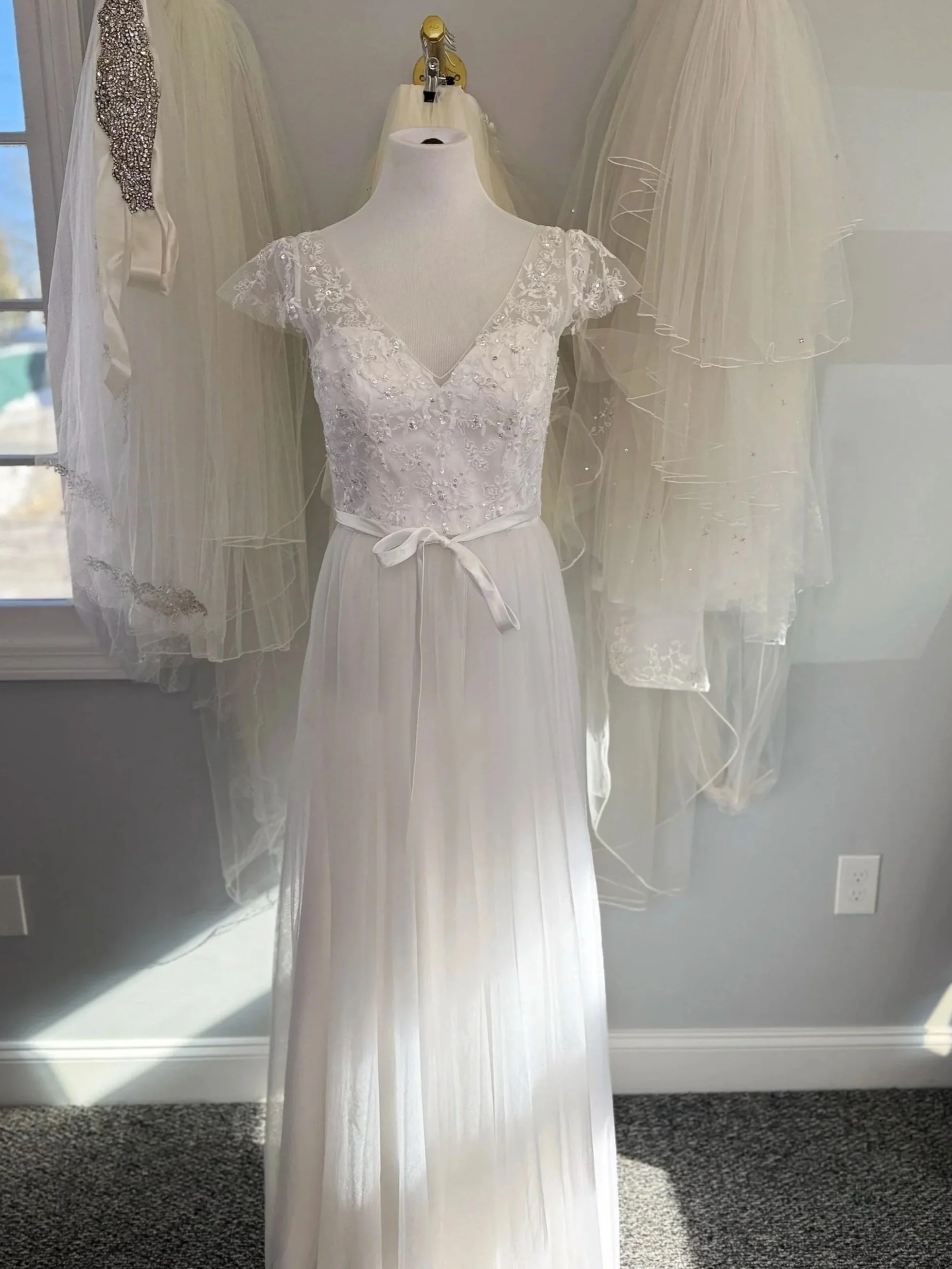 Image of cap sleeve lace bodice Morilee New York gown on a mannequin in front of veil wall.