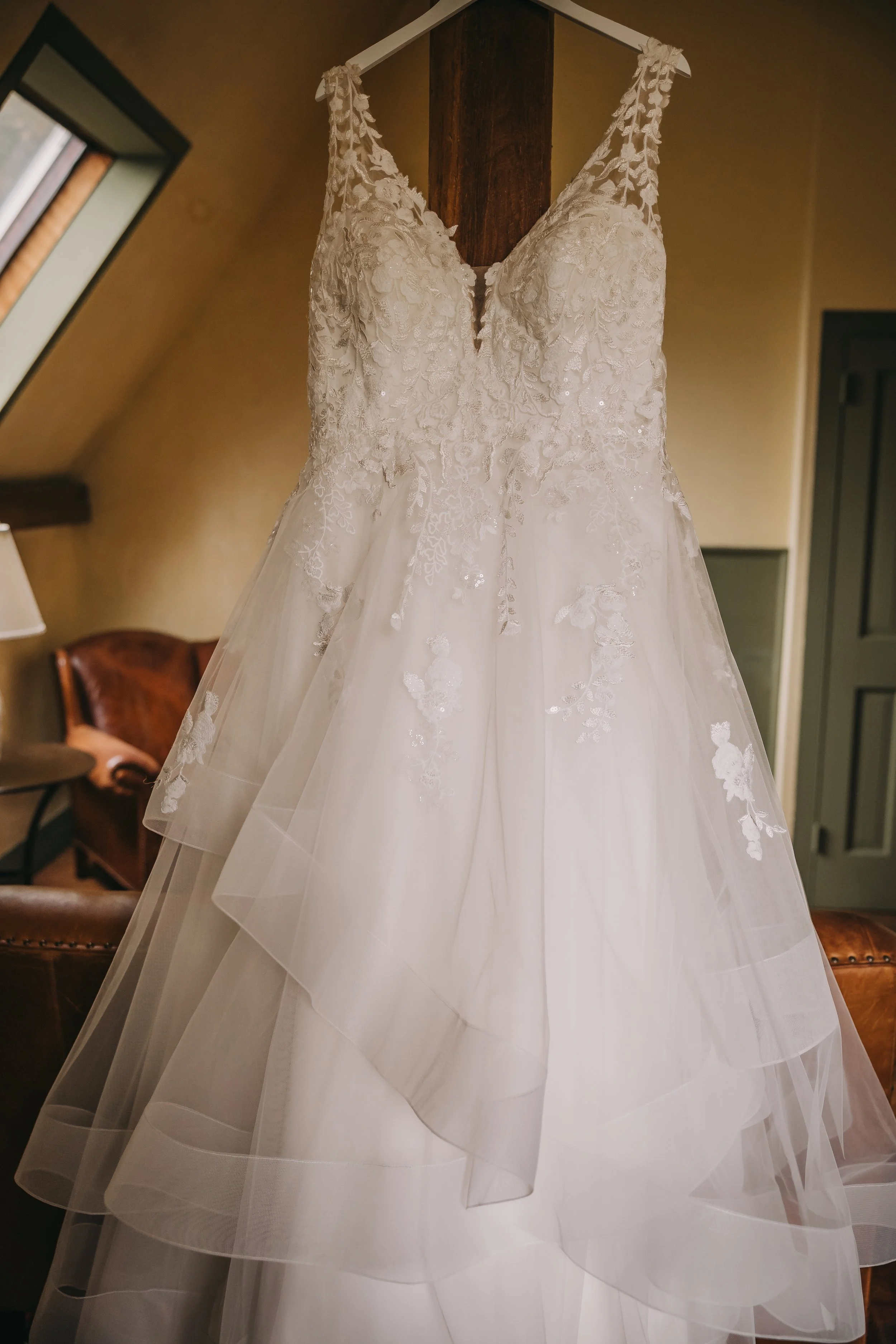 A white wedding dress with lace top and layered tulle skirt hanging on a white hanger in a room with beige walls and dark wooden furniture.