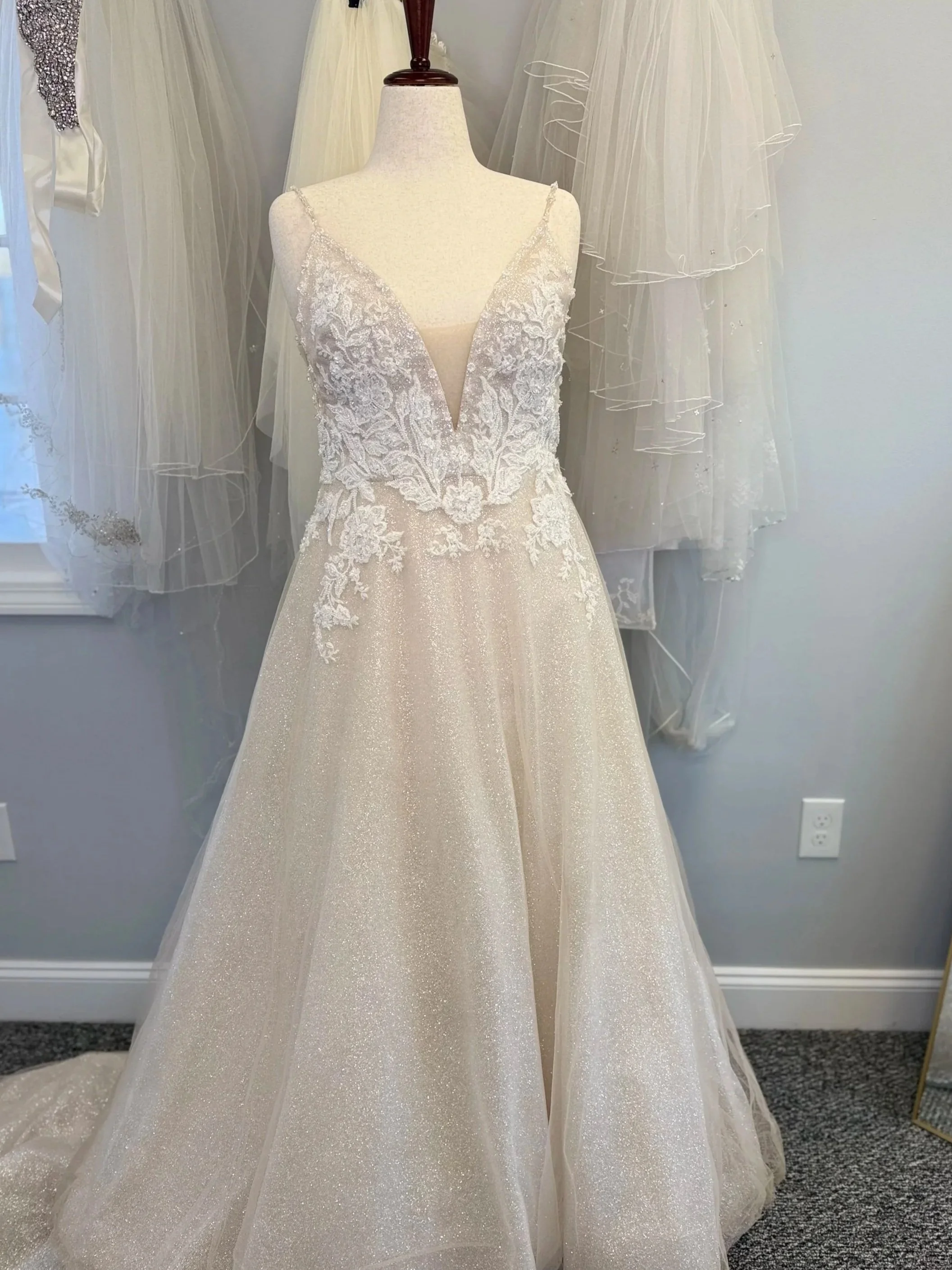 A wedding dress on a mannequin with lace and beaded details on the bodice, a deep V neckline, and a flowing, glittery skirt. Several other wedding dresses are hanging in the background.