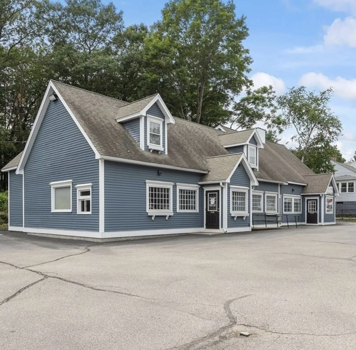 Blue building with white trim, multiple windows, and dormer windows on a cracked asphalt lot with trees in the background.