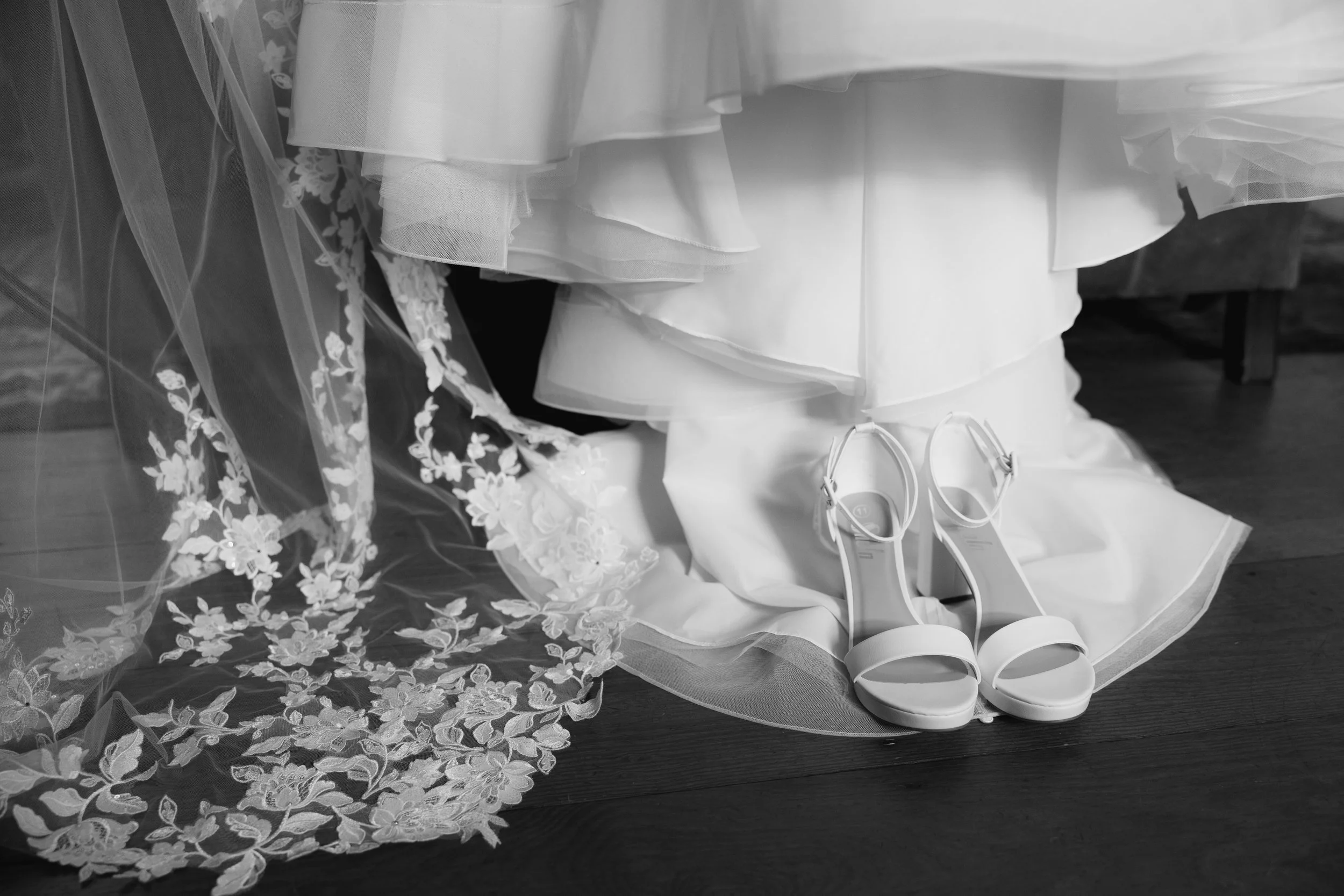 Elegant wedding dress with layered fabric and lace veil, white high-heeled shoes, and floral accessory on a wooden floor.