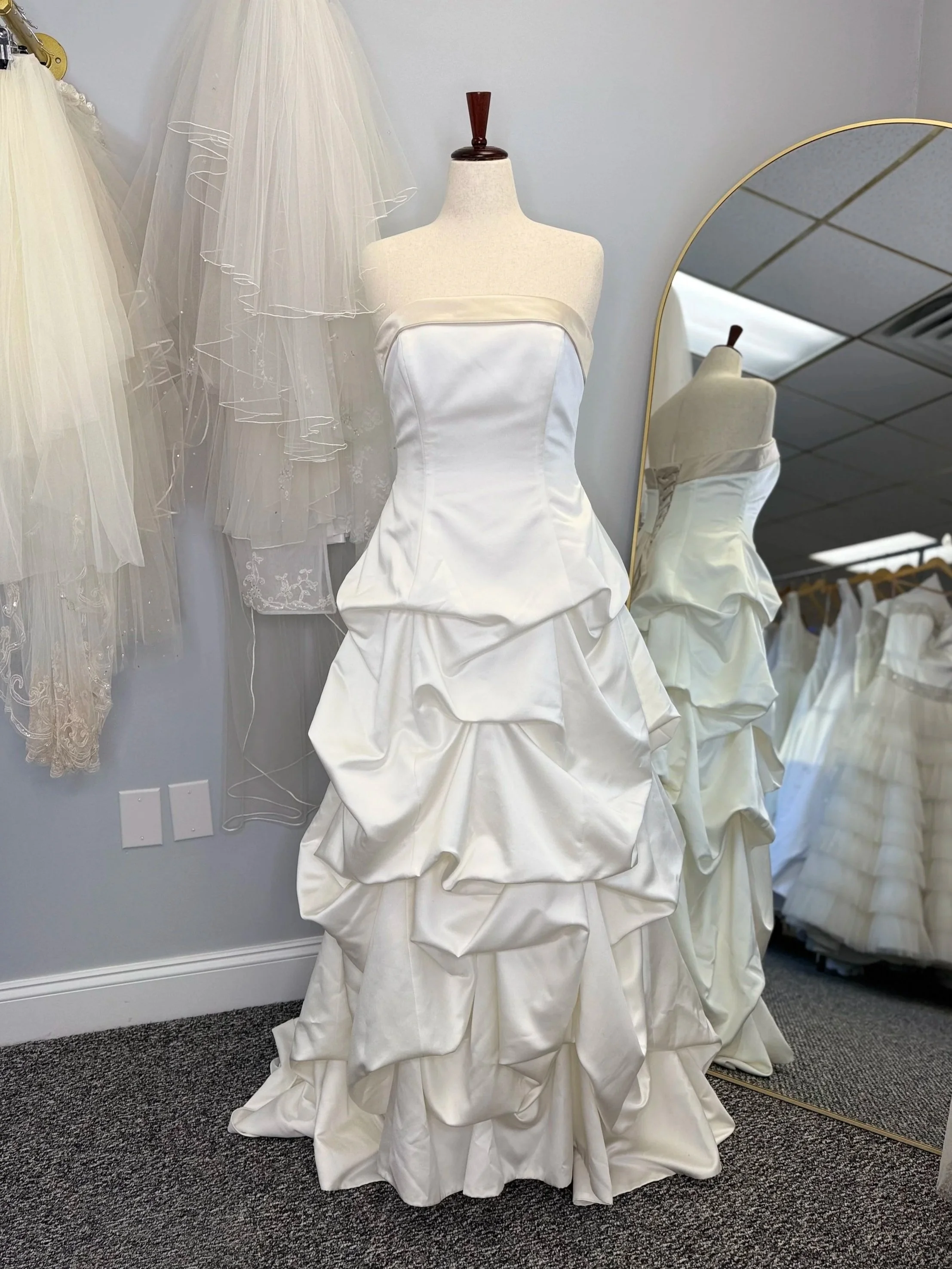 White wedding dress with a strapless neckline and layered ruffled skirt displayed on a dress form near a mirror in a bridal shop.