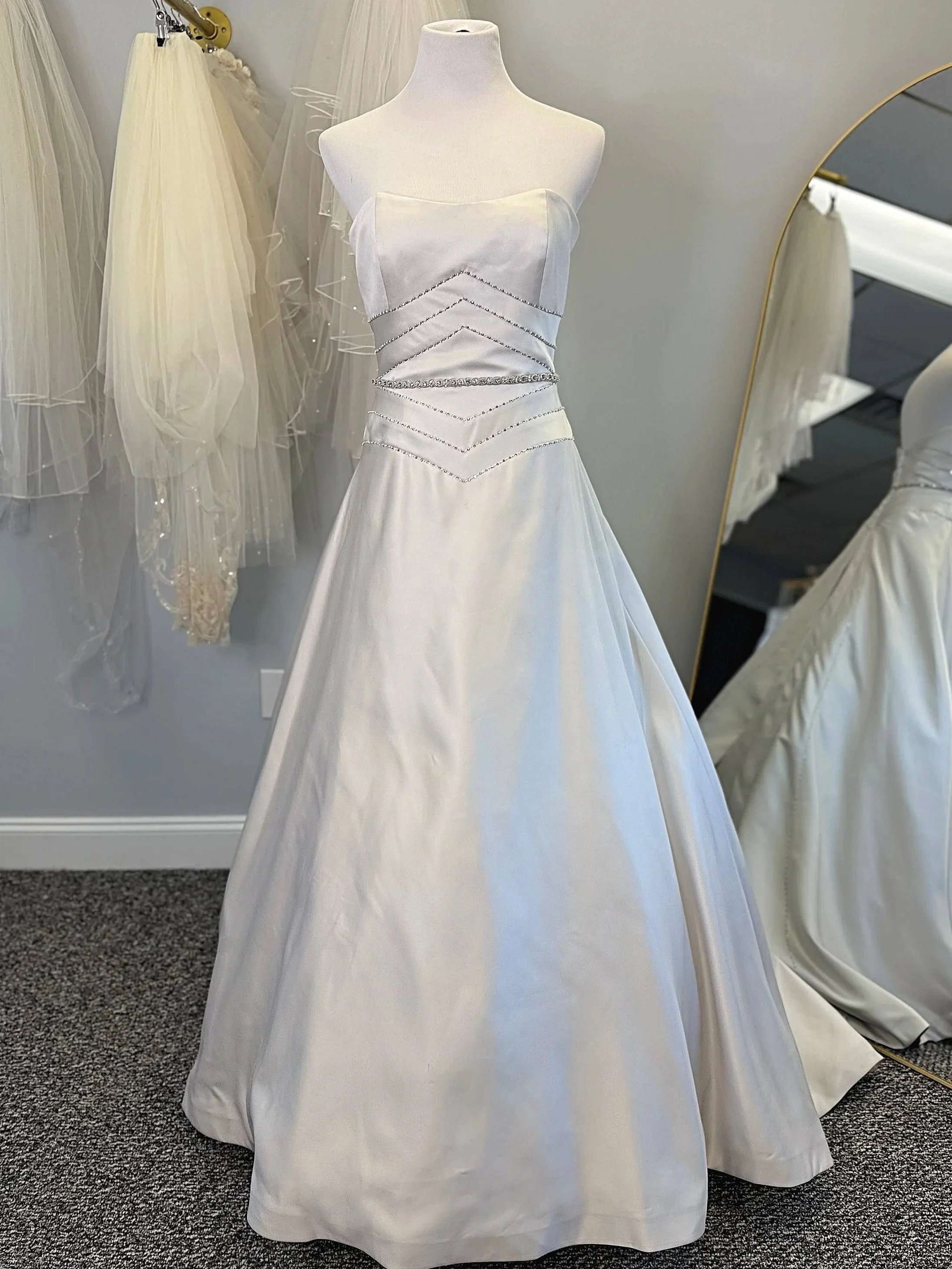 A white satin wedding gown on a mannequin, with a strapless bodice and a full skirt, adorned with silver beaded embellishments on the bust and waist area.