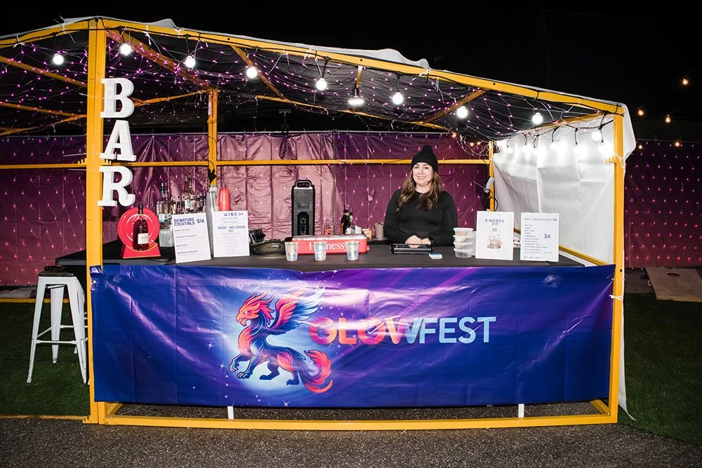 A woman standing behind a food and beverage stall at LGW Fest, decorated with string lights and a large blue banner featuring a mythical creature and the event logo. The stall has a bar sign on the left and a menu on the right.