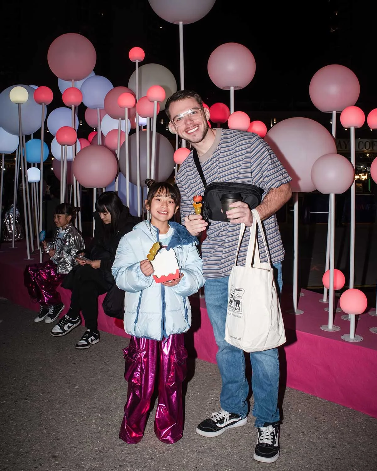 A man and a girl standing in front of a colorful display of round, glowing lights in a night setting. The girl is holding a skewer of food and a box with a cartoon face, both smiling. The man is holding a similar skewer and a drink. The girls sitting on a bench in the background are engaged with their phones.