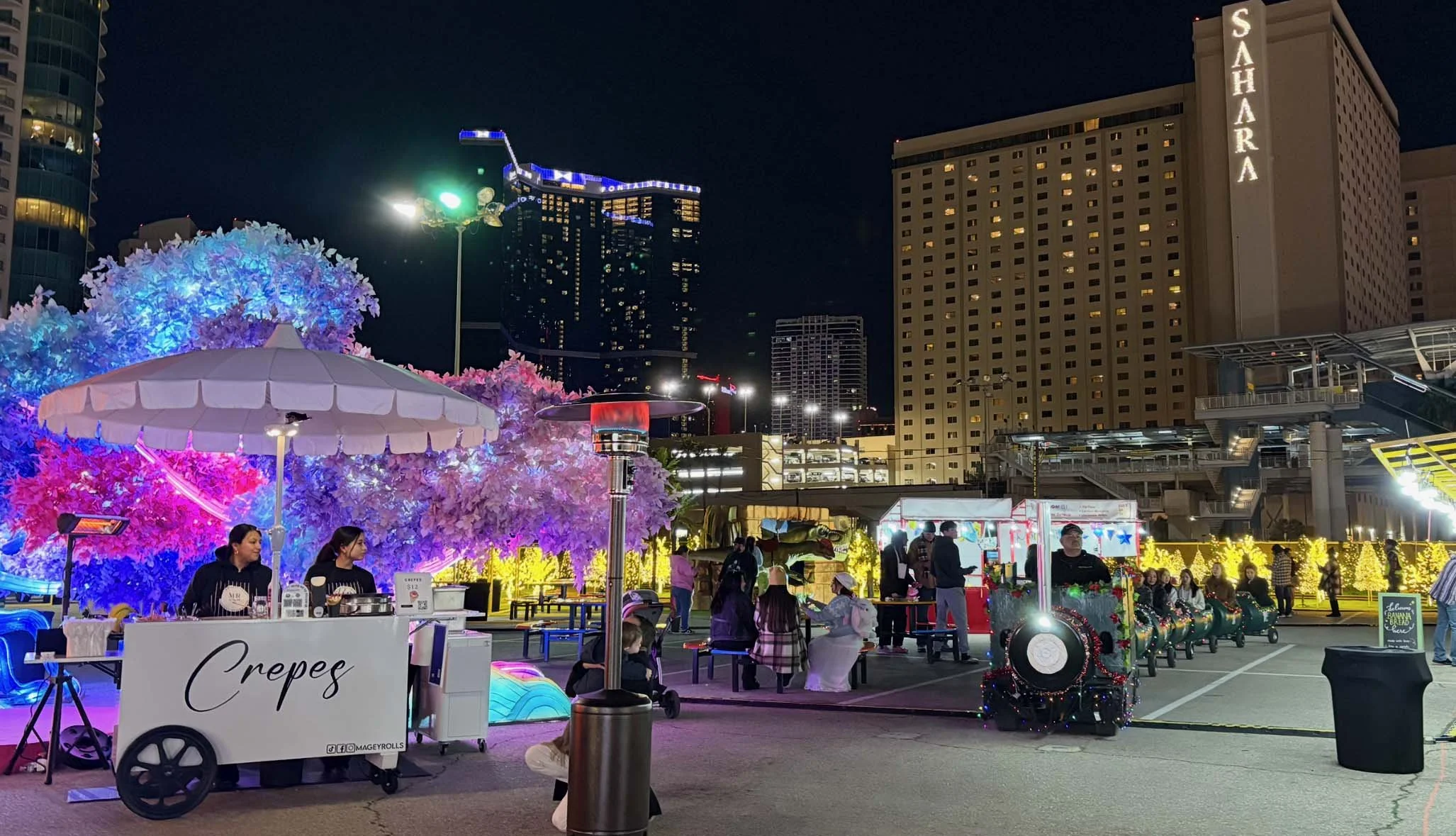 Night scene at an outdoor fair with illuminated pink and purple trees, food stall selling crepes, and people walking and sitting around decorated with holiday lights.