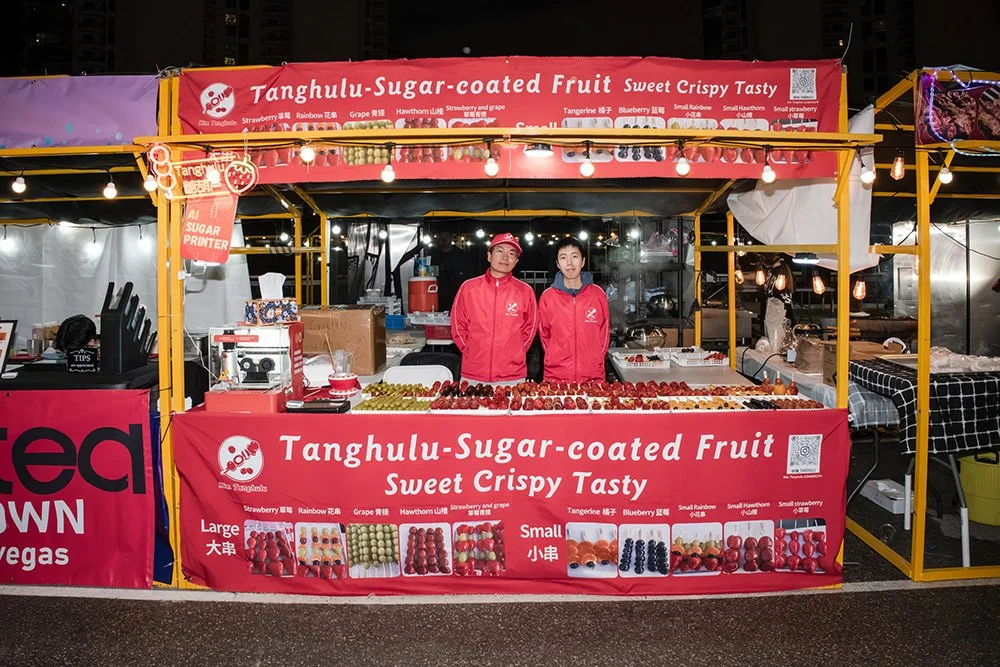 Two vendors in red jackets at a fruit stand selling sugar-coated fruit, including strawberries and grapes, with a red banner displaying product options and descriptions.