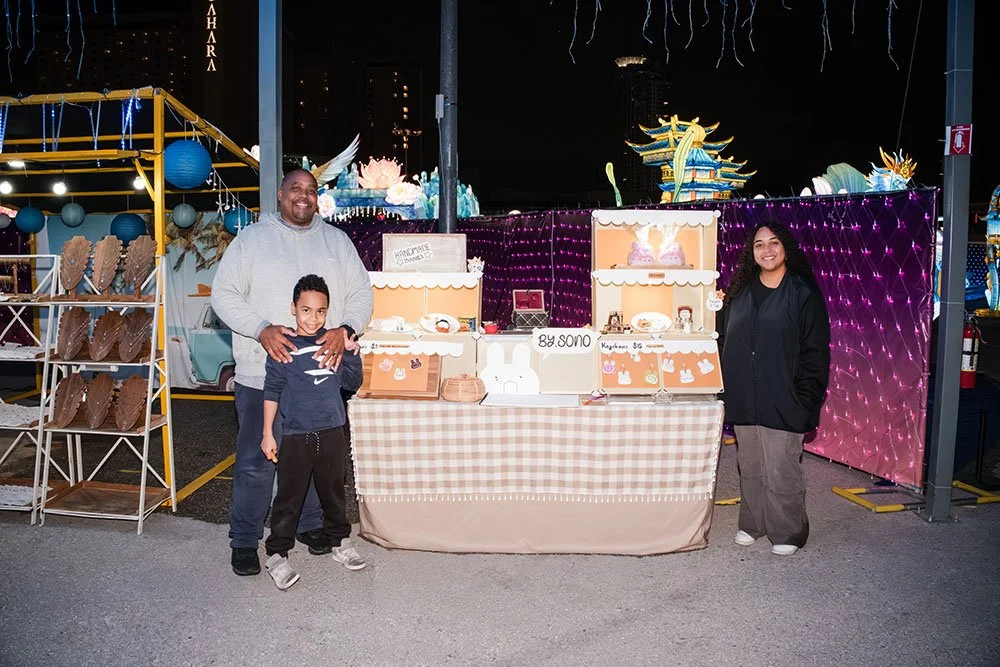 A family of three at a nighttime outdoor market stall selling handmade crafts and baked goods, with decorative lights and amusement park rides in the background.