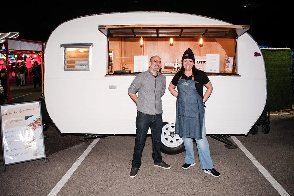 Two people standing in front of a white food truck at night, one man and one woman wearing a beanie, apron, and casual clothing, smiling at the camera.