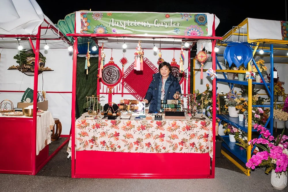 A woman standing behind a market stall decorated with jewelry, crafts, and ornaments at an outdoor night market. The stall has a sign saying 'Auspicious Garden' and is surrounded by flowers and other stalls.