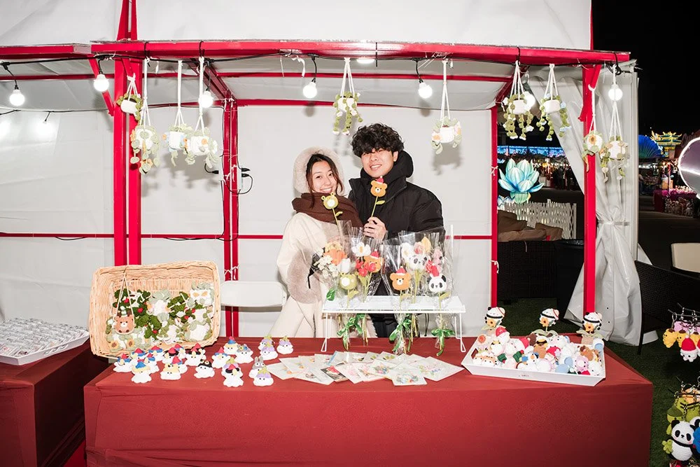 A young couple standing behind a market stall at night, decorated with plush animal and character accessories, selling felt crafts and small plush toys.