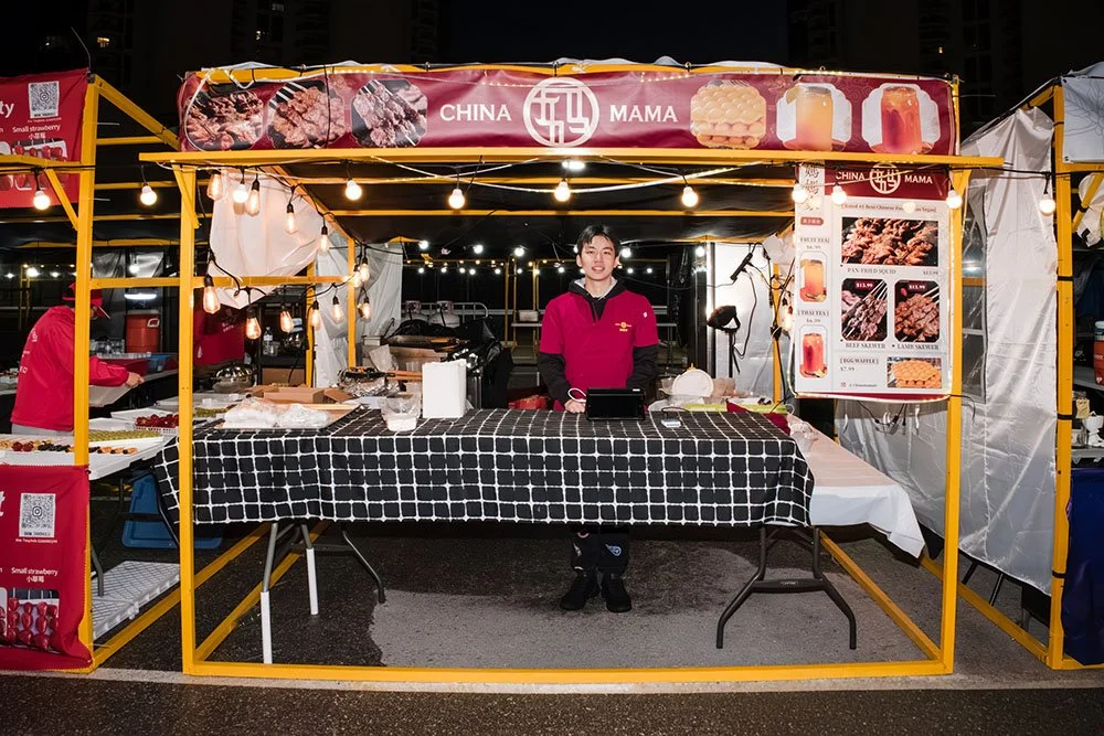 A food stall named 'China Mama' at night, with a young person standing behind a checkered table. The stall is decorated with string lights and has a menu board with Chinese food items and drinks displayed.
