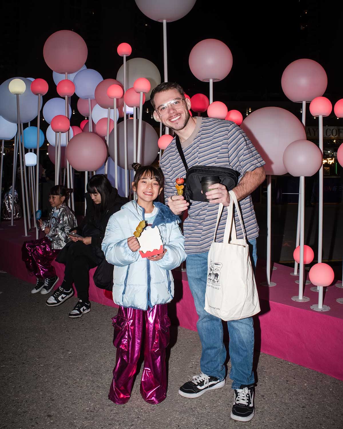 A man and a girl smiling and holding food stand in front of pink, white, and blue globe-shaped lights at night, with two seated children behind them.