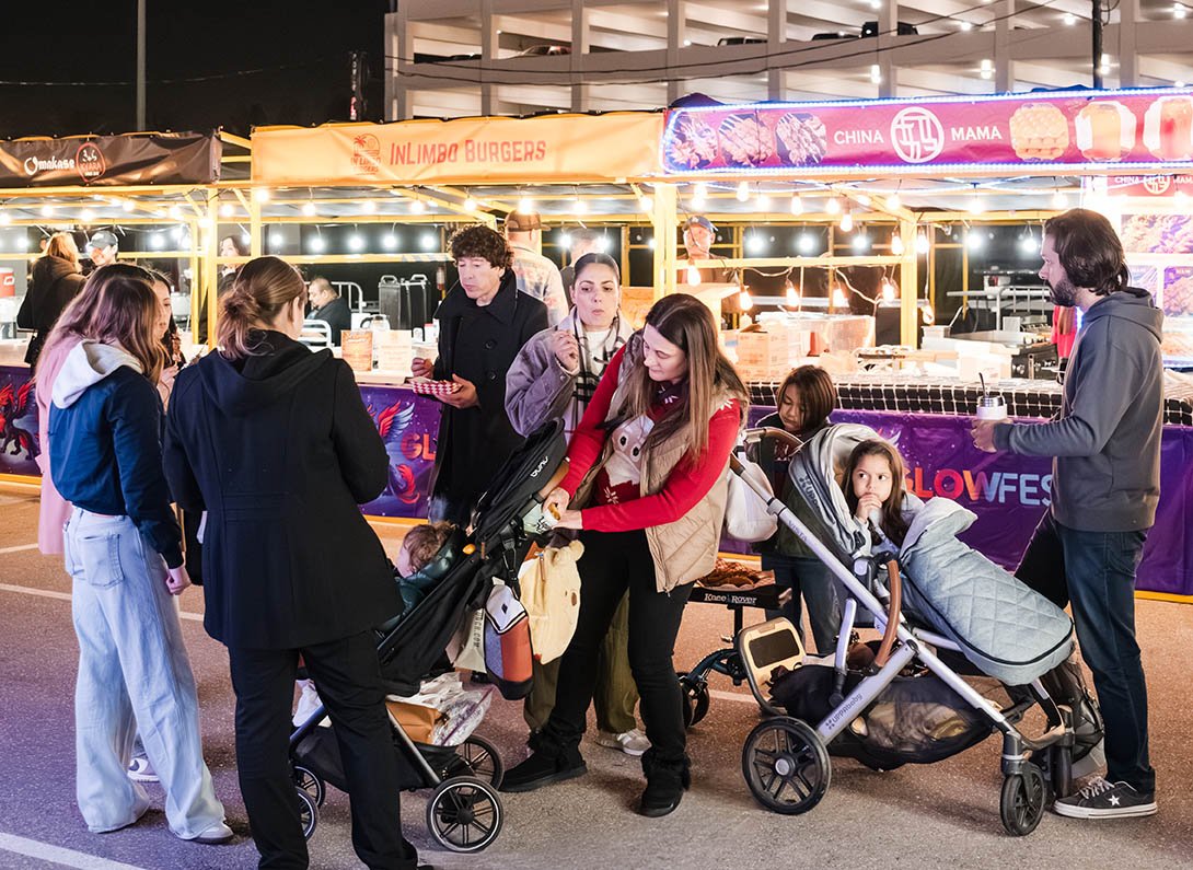 A group of people including adults and children waiting in line at a night market or food festival, with food stalls and lights in the background.