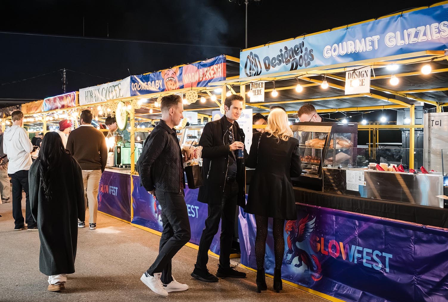 People standing in line at a food stall at night, with bright lights and colorful banners advertising gourmet grilled dogs and other treats at a fair or festival.