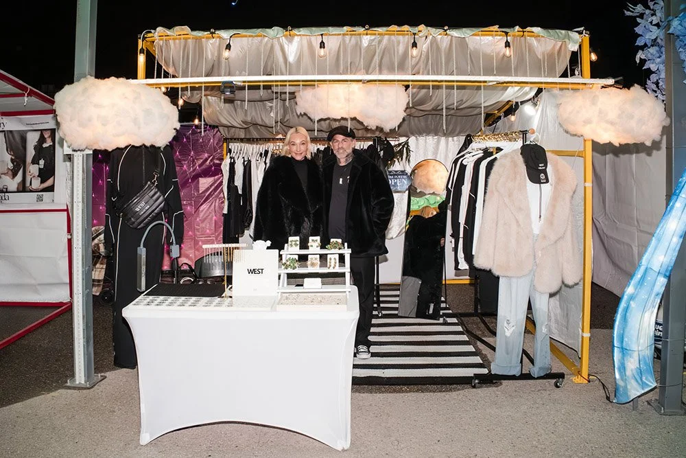 A booth at an outdoor night market with clothing displays, two people standing behind the counter, and decorative cloud-shaped lights.