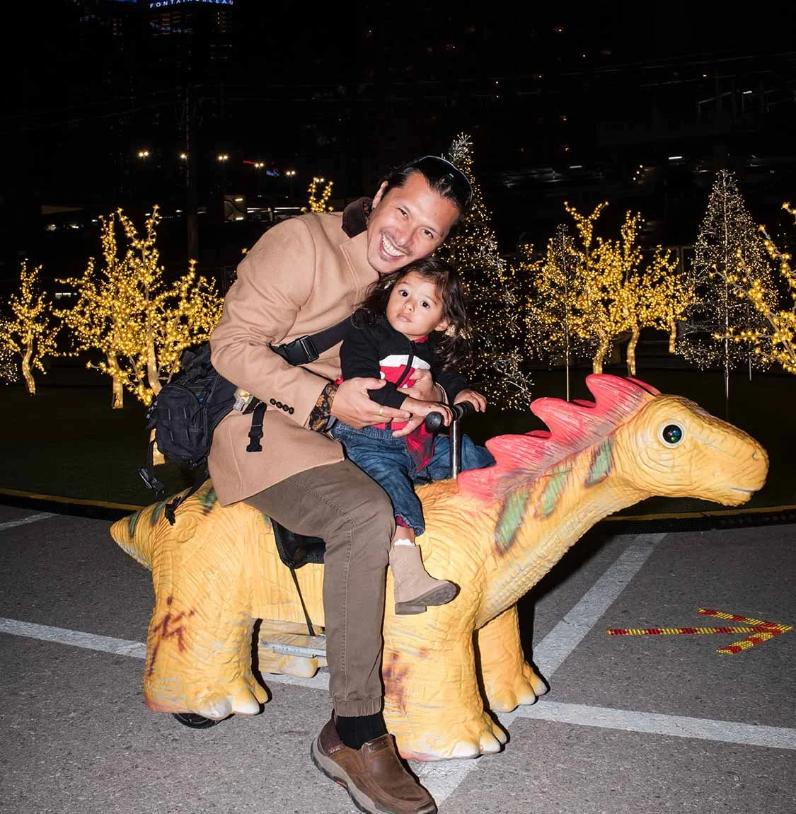A man and a young girl ride a dinosaur-shaped playground sculpture outdoors at night, with decorative lights and trees in the background.