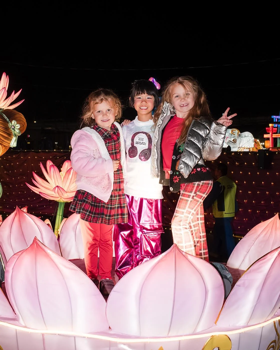 Four young girls dressed in colorful outfits standing inside a large, illuminated pink flower float at a nighttime festival or parade.