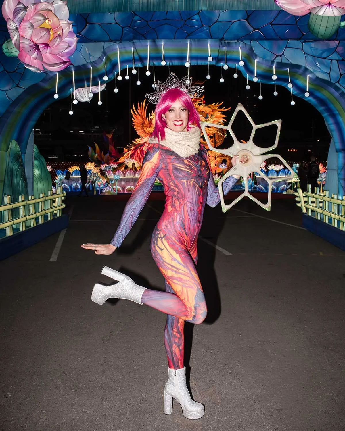 Person in a colorful, artistic costume with pink hair, standing on one leg and holding a star-shaped prop, with illuminated flower and nature-themed decorations in the background.