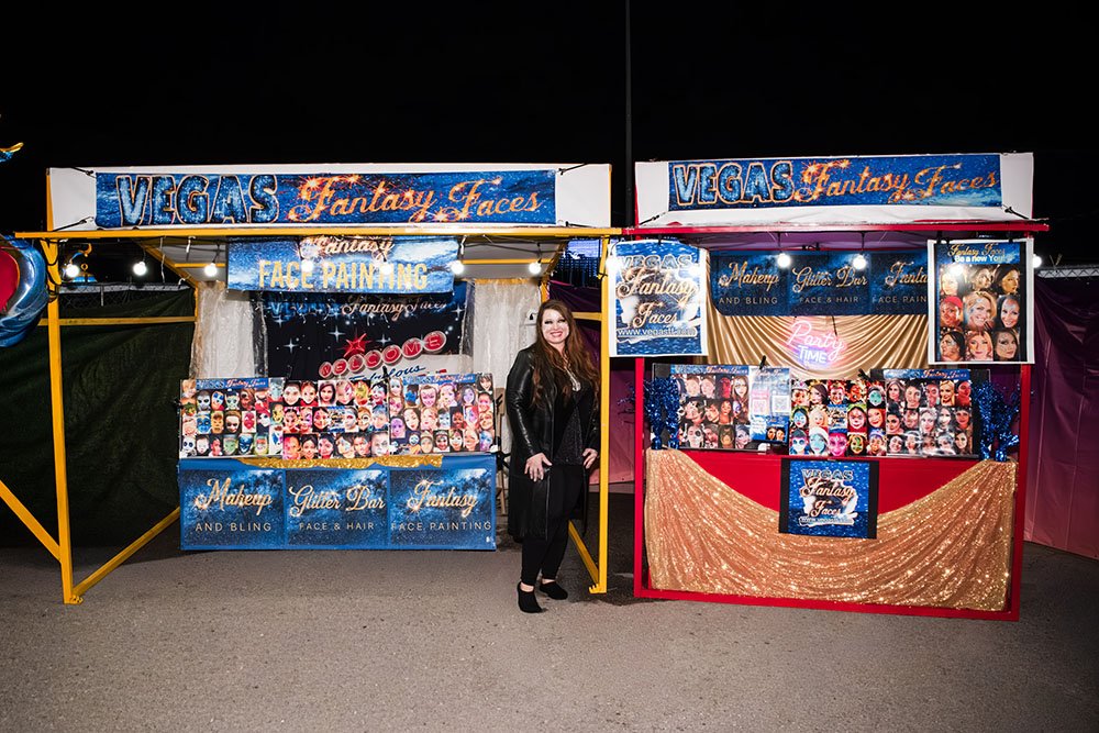 Woman standing between two booth setups at Vegas Fantasy Faces, offering face painting, makeup, glitter bar, and face and hair bling, with colorful signs and display of face masks and painted faces.
