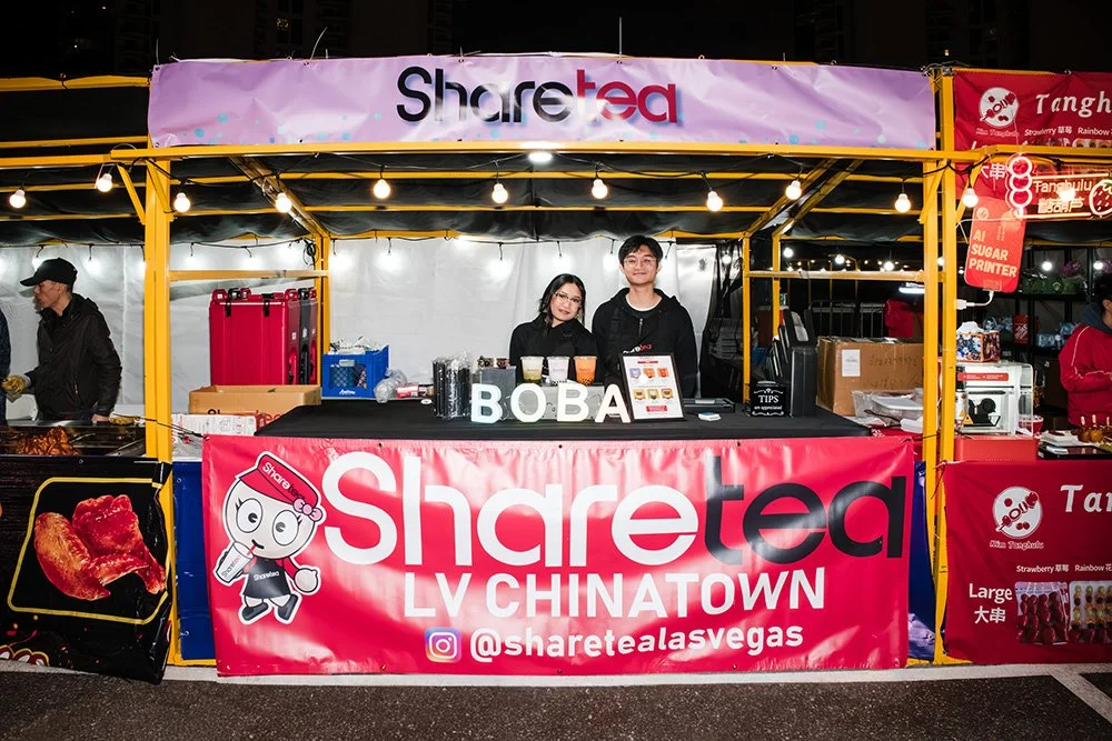A food stall at a night market named Sharetea, located in LV Chinatown. Two staff members stand behind the counter with a pink banner displaying the store's name and social media handle. The stall offers Boba drinks, as indicated by the letters on th