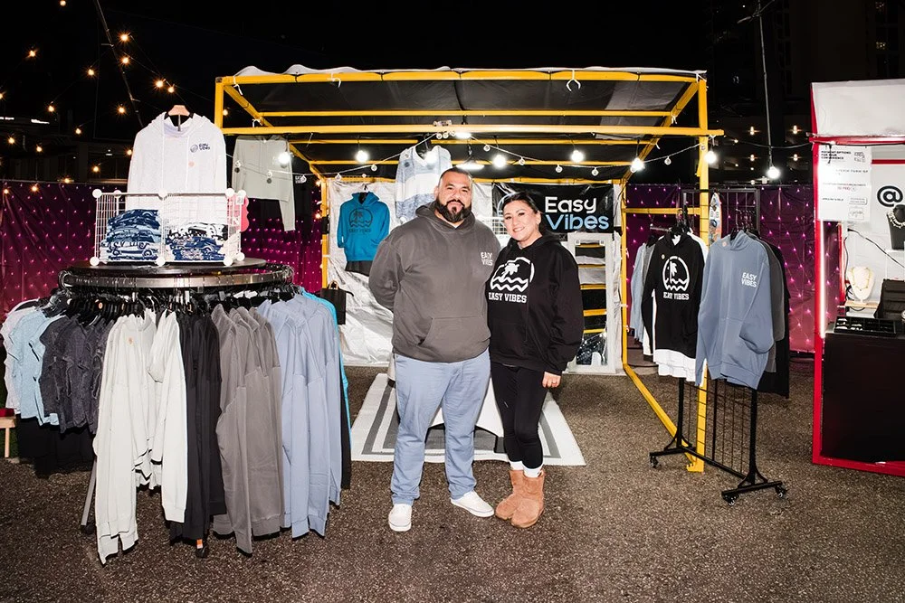 Two people standing inside a clothing booth with racks of hoodies and shirts at an outdoor market or event at night.
