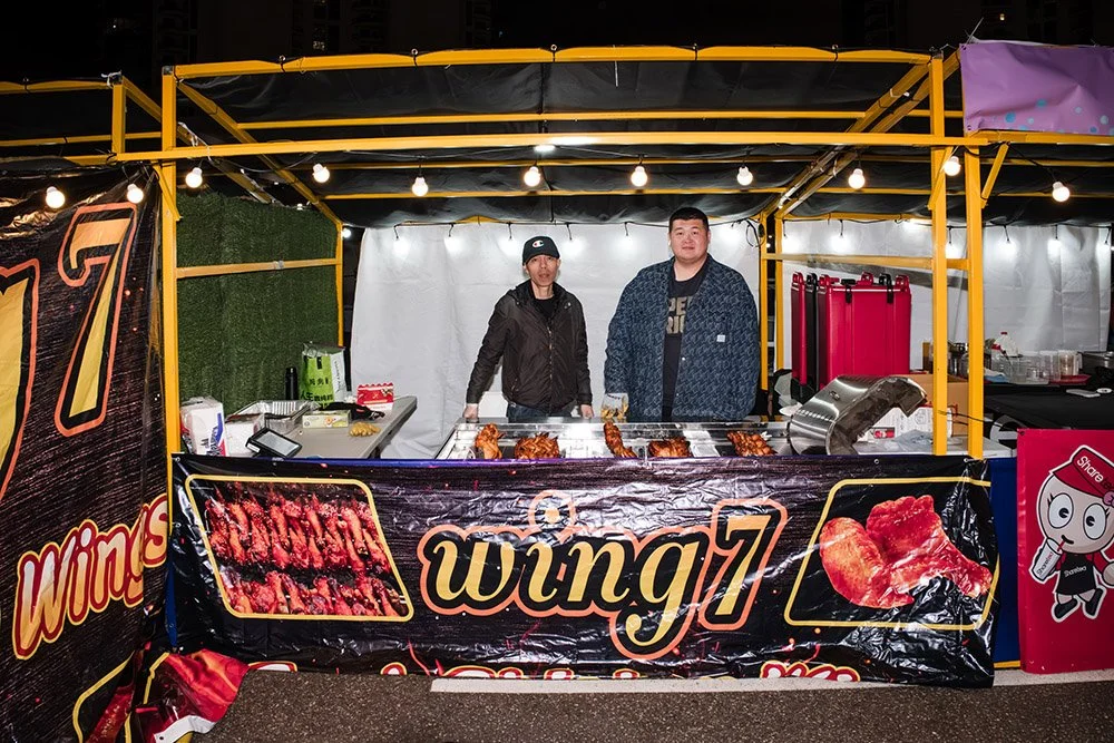 Two men standing behind a barbecue grill booth at night. The booth is decorated with a sign showing grilled meat and the text 'wung7'. There is string lighting overhead, and the background features city buildings.