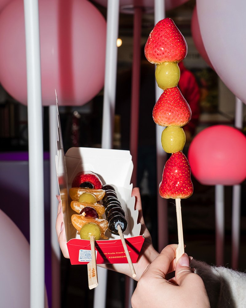 Person holding a skewer with strawberries and green grapes, and a box of assorted fruit skewers in the background, with pink and purple balloons in a festive setting.