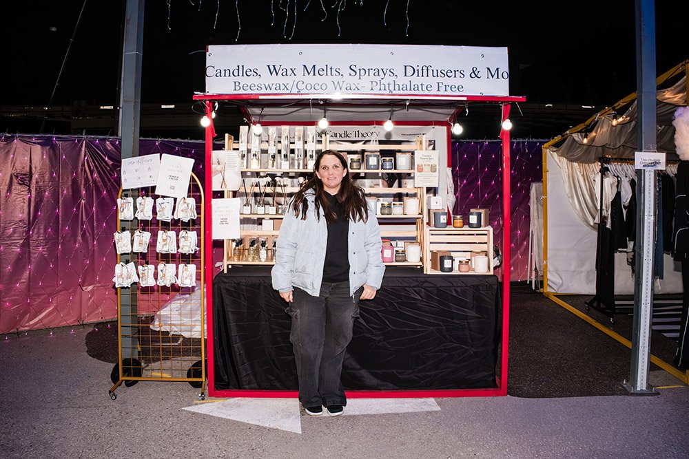 A woman standing in front of a candle and wax melts booth at a market, with a sign advertising candles, wax melts, sprays, diffusers, and more, made from beeswax and coconut wax. The booth has shelves with various scented candles.