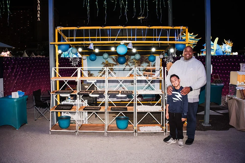 A man and a boy standing together at a holiday or carnival event, in front of a display with empty shelves and seasonal decorations, illuminated by string lights and blue paper lanterns.