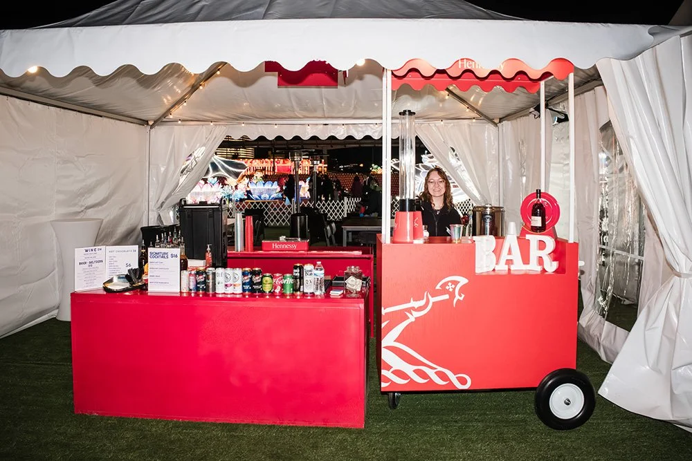 A woman standing behind a red bar cart inside a white tent at night, with various drinks, cups, and a menu on the cart. The word 'BAR' is displayed on the cart, and there are decorative elements on the front.