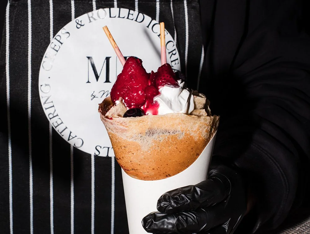 A person in a black glove holding a crepe cone filled with whipped cream, strawberries, and blueberries.