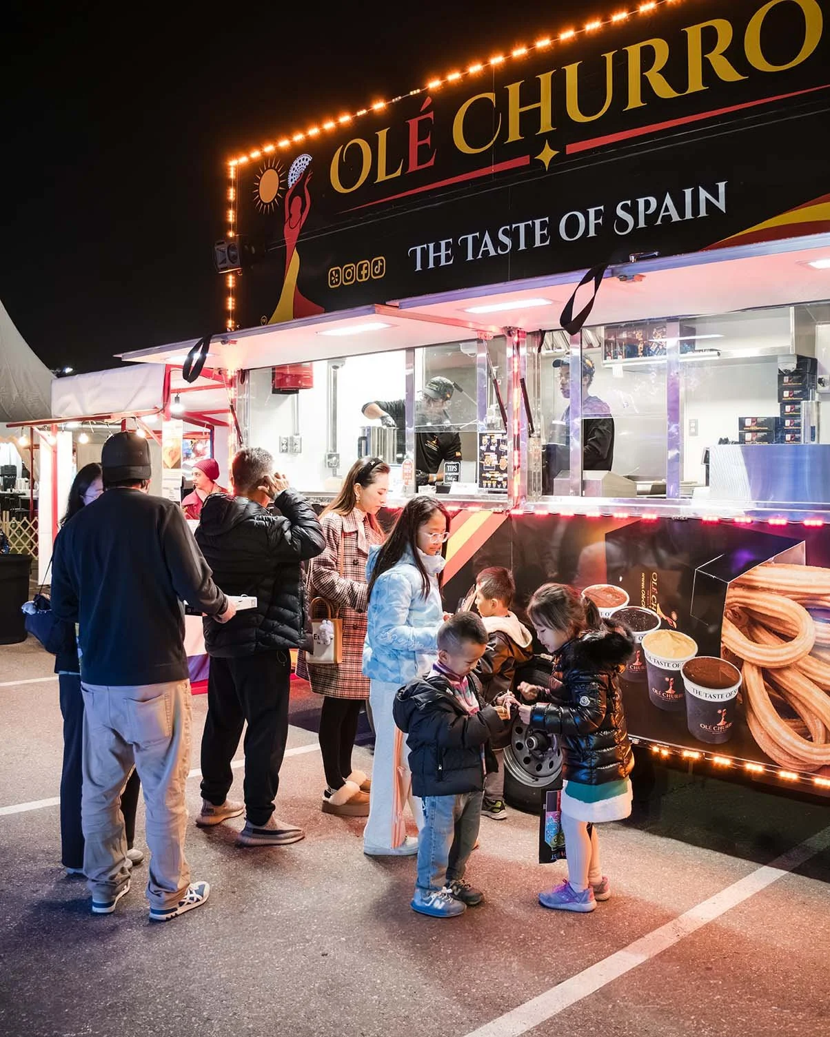 People in line at a churro food stand at night, with a bright sign that reads 'Olé Churro' and 'The Taste of Spain', serving desserts through a window in a festive outdoor setting.