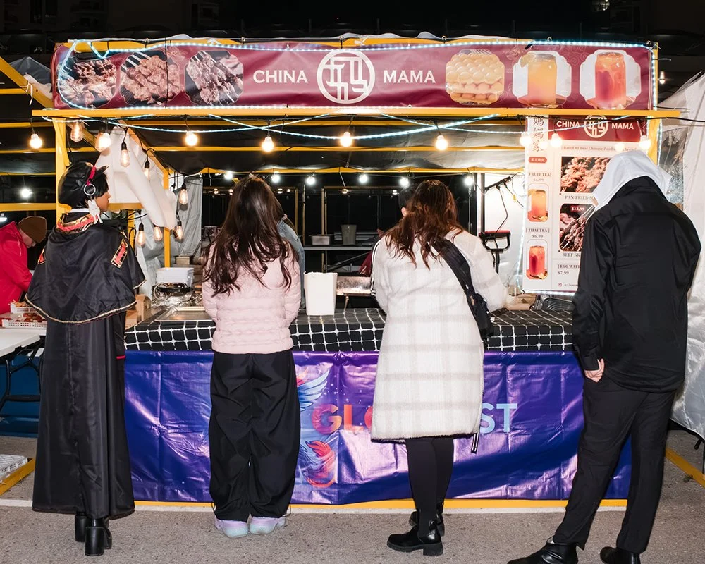 Four people standing in front of a Chinese food stall at a night market, with a banner displaying Chinese dishes and beverages.
