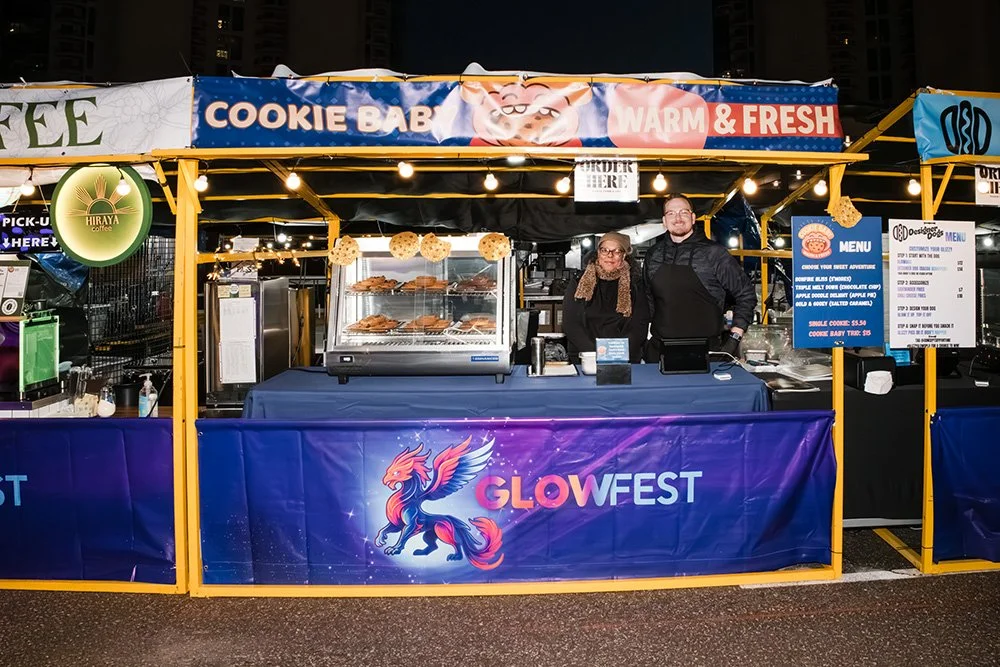 A food stall at Glow Fest selling baked goods like cookies and pastries. Two people stand behind the counter in front of a display case with baked items. The stall is decorated with a blue and purple banner with a phoenix logo and the text "Glow Fest