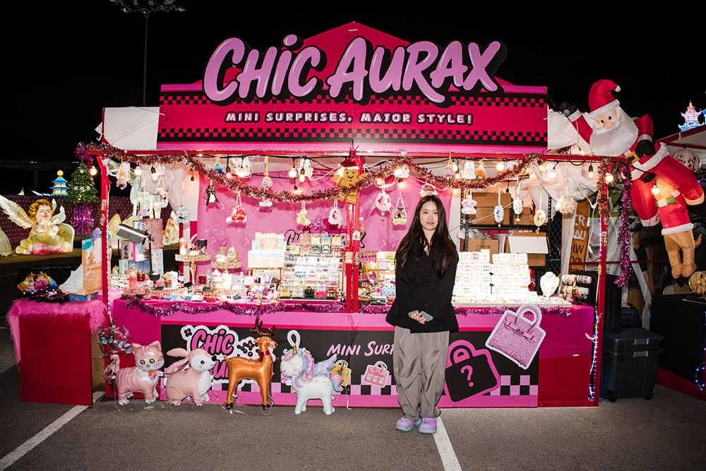 A young woman with long dark hair stands in front of a colorful market stall called "Chic Aurax" at night. The stall is decorated with pink and purple lighting, plush toys like unicorns and a reindeer, and holiday decorations, including a Santa figur