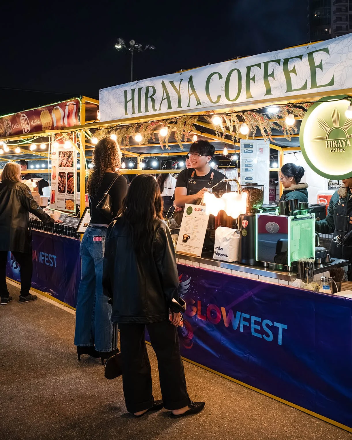 Night market food stall named 'Hiraya Coffee' with customers ordering and a bartender serving coffee, illuminated by string lights.