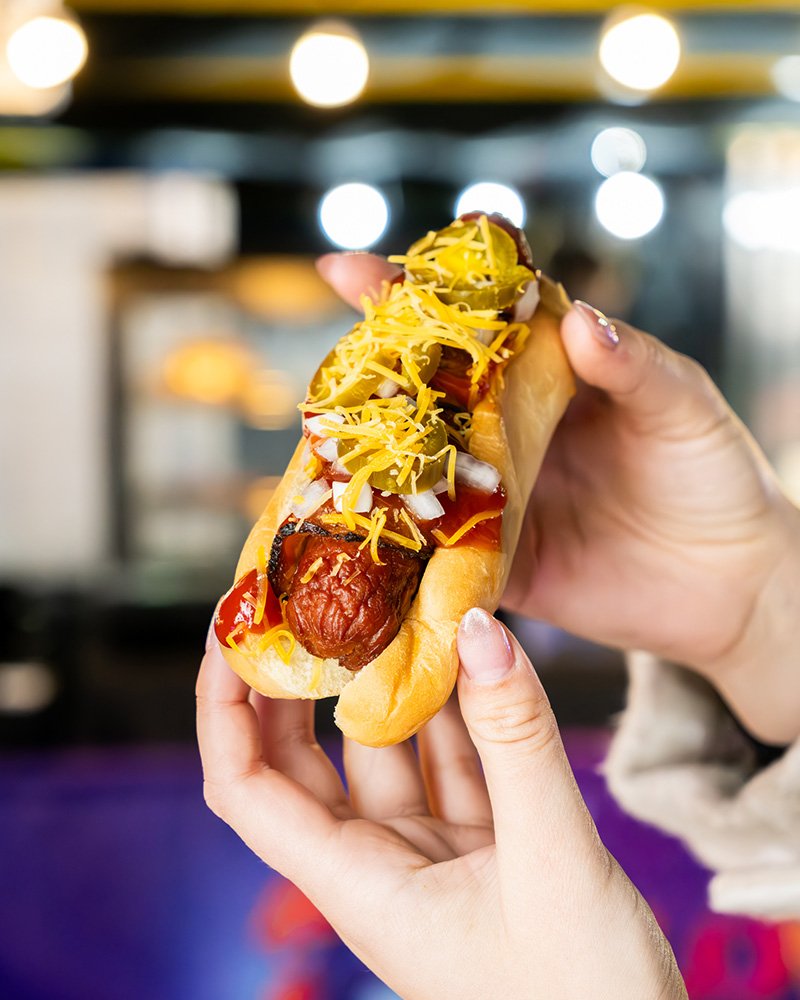 Hot dog with mustard, shredded cheese, and chopped onions in a bun, held by a person's hands.