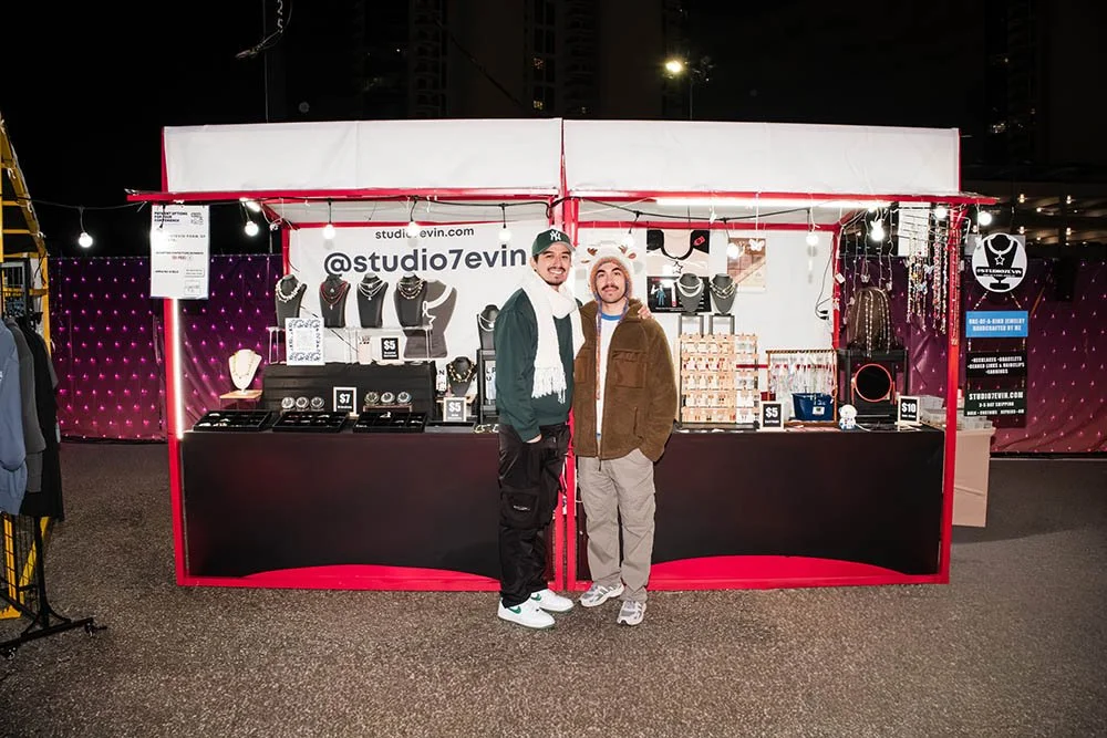 Two men smiling at a jewelry stand at night. The stand offers necklaces, rings, and earrings for sale. One man wears a New York Yankees baseball cap and scarf, the other wears a fluffy hat and brown jacket.