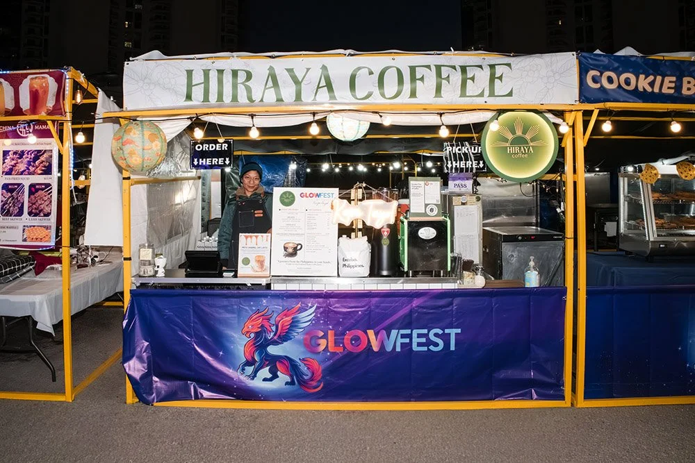 A booth at an outdoor festival with a sign that reads 'Hiraya Coffee' and a glowing circular sign with the same name. The booth is decorated with string lights and has a banner at the front with a colorful dragon and the text 'GLOWFEST.' A person is 