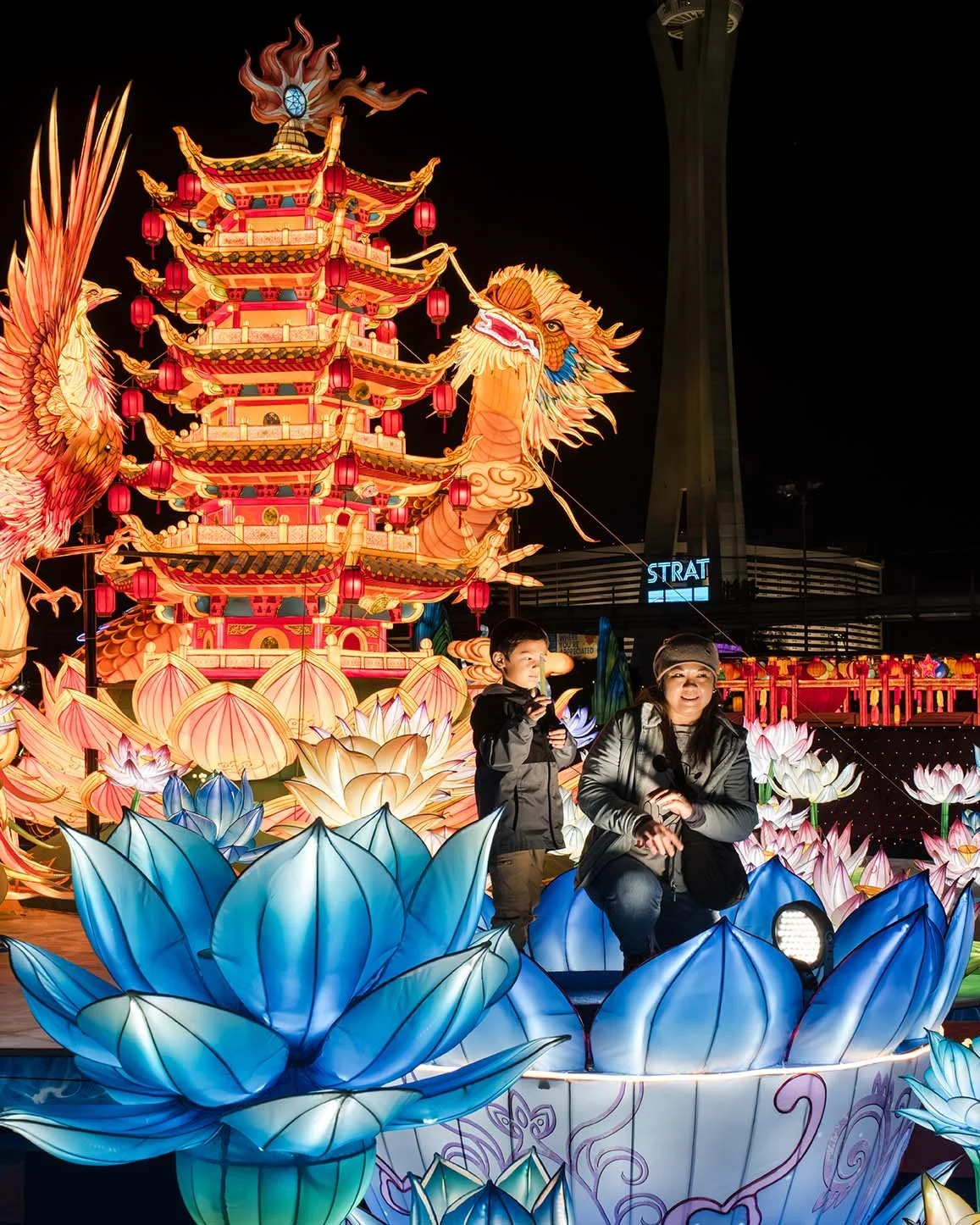 People riding a colorful illuminated lantern float featuring a multi-tiered pagoda, a phoenix, and lotus flower designs at night.