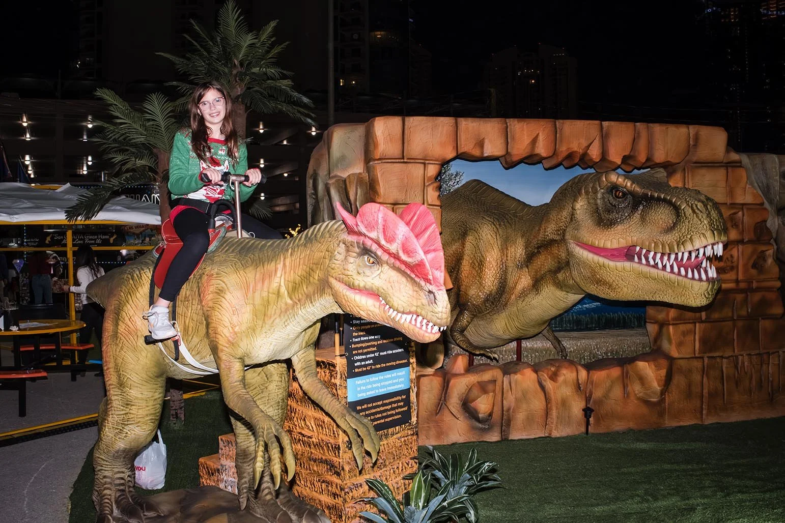 A girl riding a dinosaur-themed amusement ride at night, with a large dinosaur backdrop and artificial greenery around.