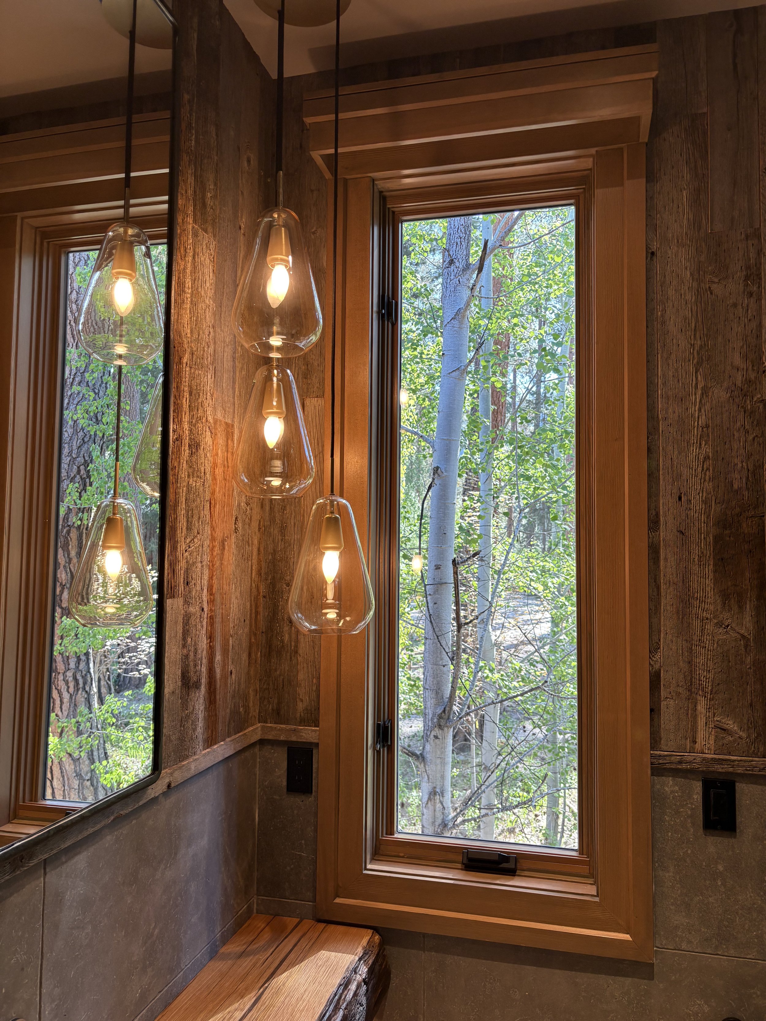 Interior of a room with tall wooden-framed windows showing a view of trees outside. Modern pendant lights with glass covers hang from the ceiling, casting light on a rustic wooden bench below. The walls are made of textured wood and stone materials.