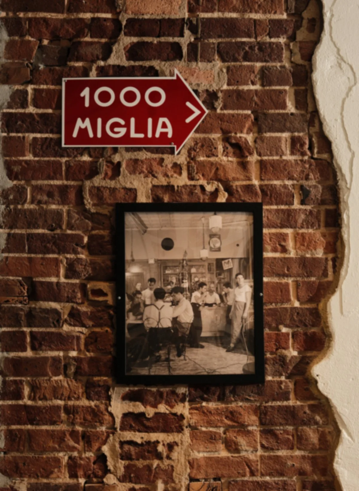 A brick wall with a red and white sign pointing to the right that reads '1000 MIGLIA' and an old black and white photograph of people inside a restaurant or bar.