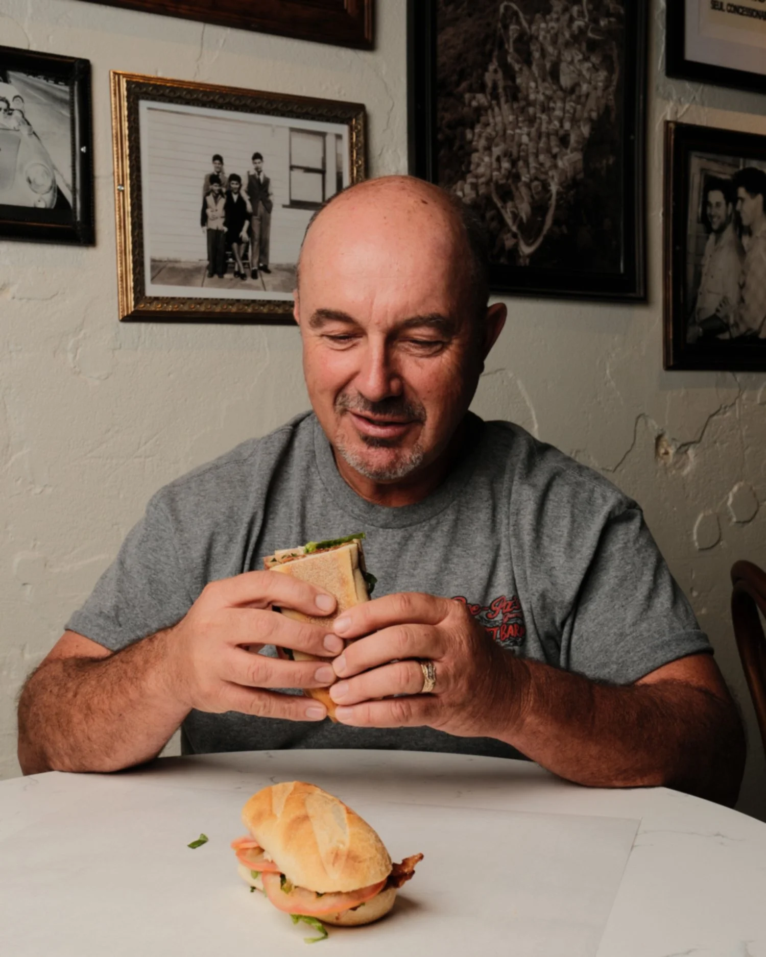 A man sitting at a table, holding a sandwich, with a small sandwich on the table in front of him, against a wall decorated with framed photographs.