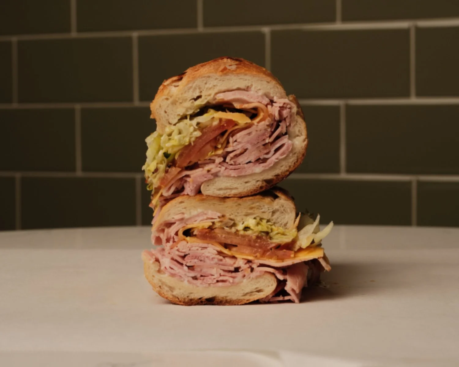 Stacked halved sandwiches with turkey, ham, cheese, lettuce, and tomato on a white surface with dark tiled wall in background.