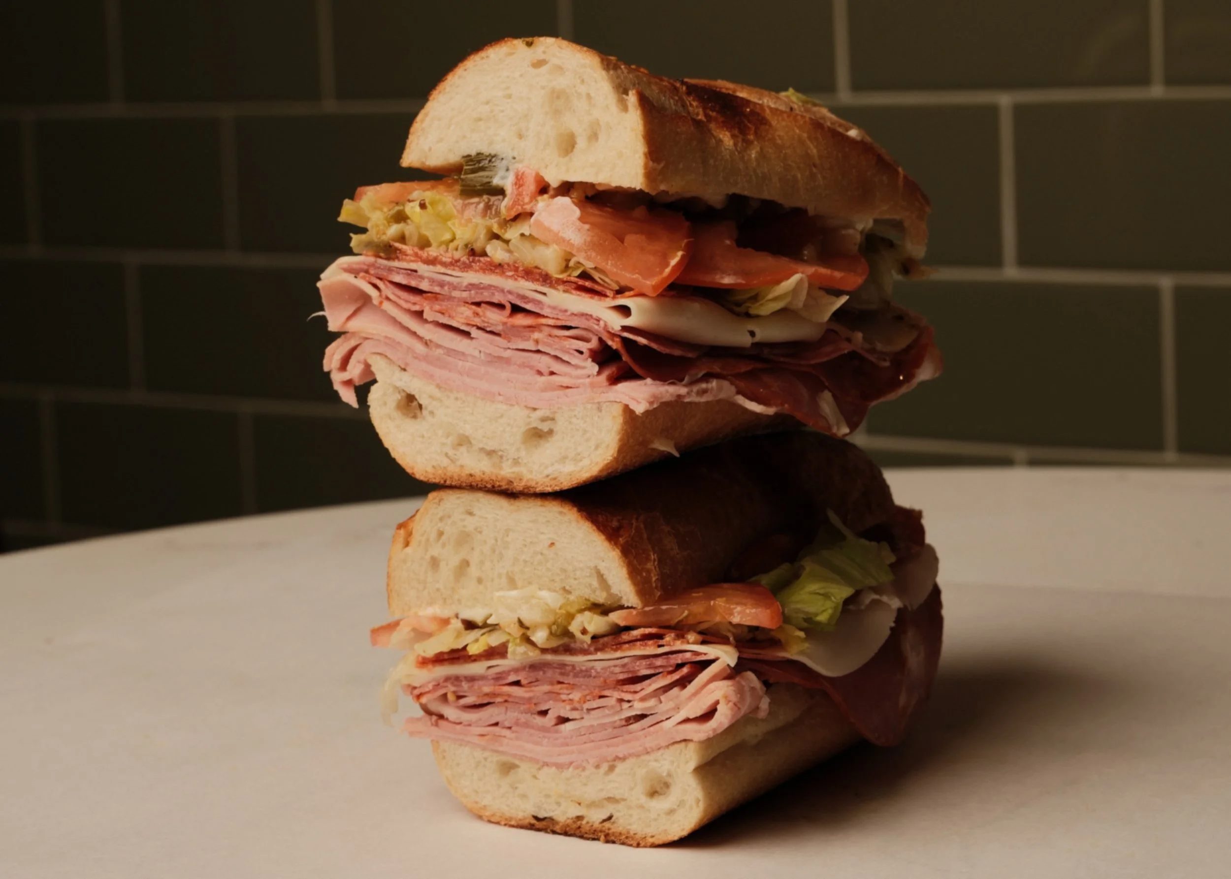 Stacked deli sandwiches with turkey, ham, salami, lettuce, tomato, and pickles on white bread and a baguette, on a white surface with a dark tiled background.