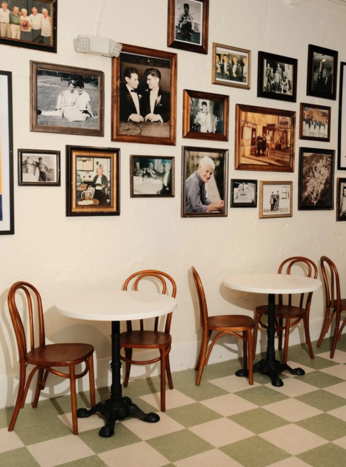 Interior of a cafe with two white round tables and wooden chairs, decorated with a collage of framed black and white and color photographs on the wall.